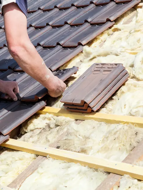 Hände eines Dachdeckers legen neue Dachziegel auf die Lattung über der Dämmung.