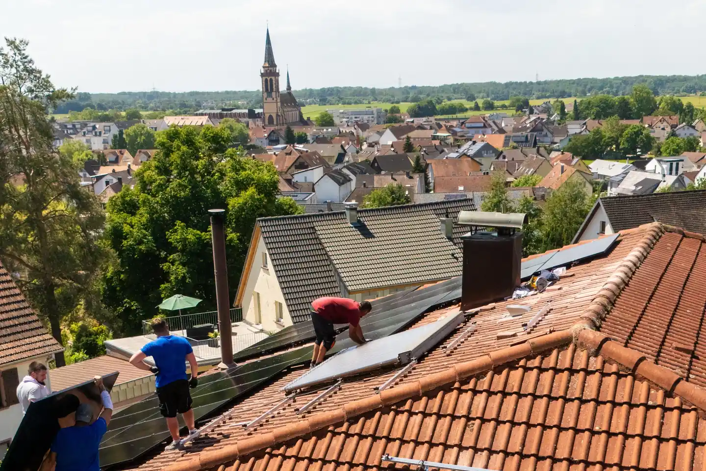Drohnenblick über ein Wohngebiet mit Kirchturm im Hintergrund: Ein Montageteam verlegt Solarmodule auf einem Ziegeldach.