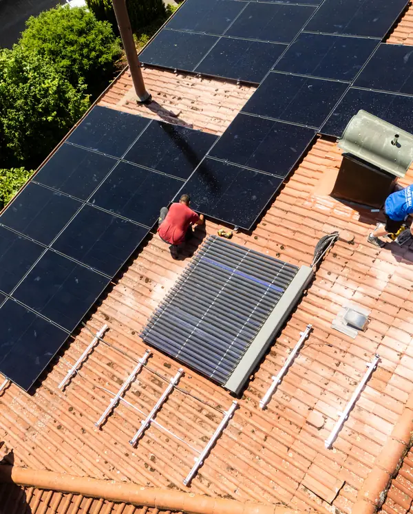 Blick von oben auf ein Ziegeldach: Zwei Monteure setzen schwarze Solarmodule neben einen Dachkollektor.