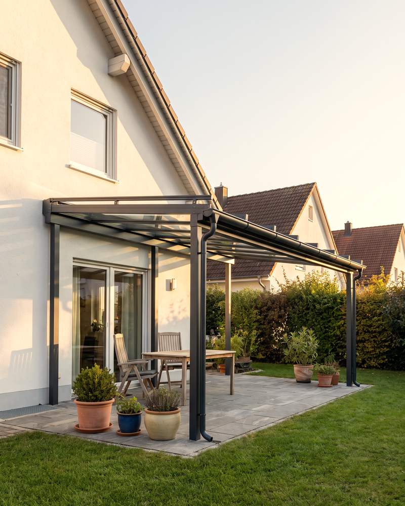 Anthrazitfarbene Terrassenüberdachung mit Glasdach an einem weißen Einfamilienhaus, darunter Gartentisch und Stühle.